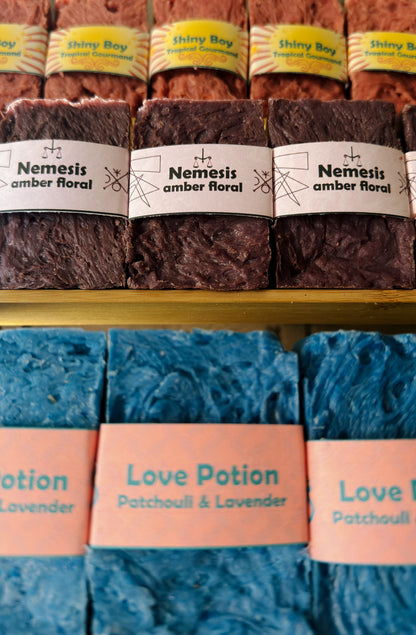 Bouncing Studio handmade bar soaps displayed on shelf including pink Shiny Boy Tropical Gourmand top row purple Nemesis Amber Floral middle row and blue Love Potion Patchouli and Lavender bottom row