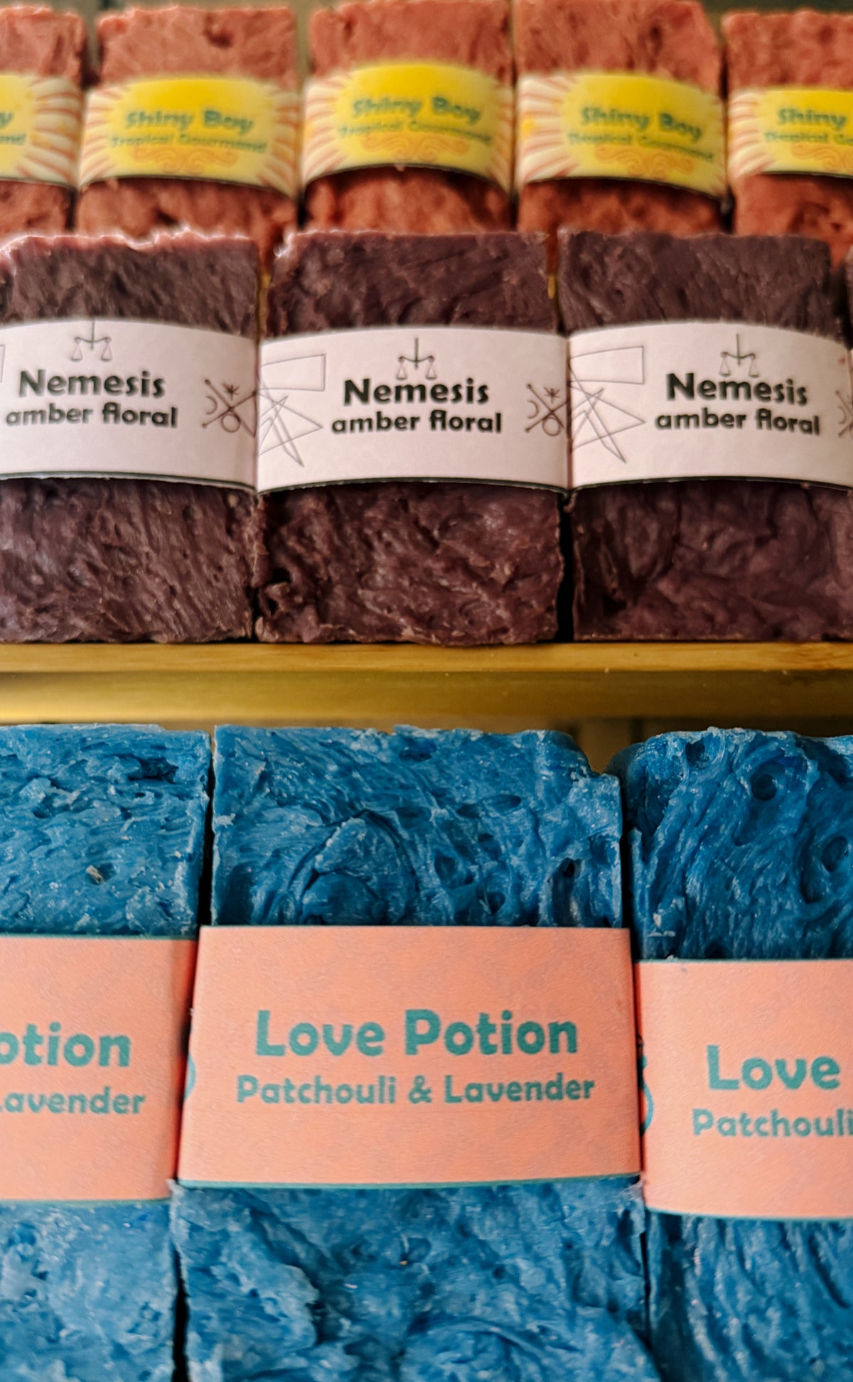 Bouncing Studio handmade bar soaps displayed on shelf including pink Shiny Boy Tropical Gourmand top row purple Nemesis Amber Floral middle row and blue Love Potion Patchouli and Lavender bottom row