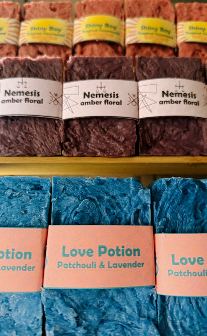 Bouncing Studio handmade bar soaps displayed on shelf including pink Shiny Boy Tropical Gourmand top row purple Nemesis Amber Floral middle row and blue Love Potion Patchouli and Lavender bottom row