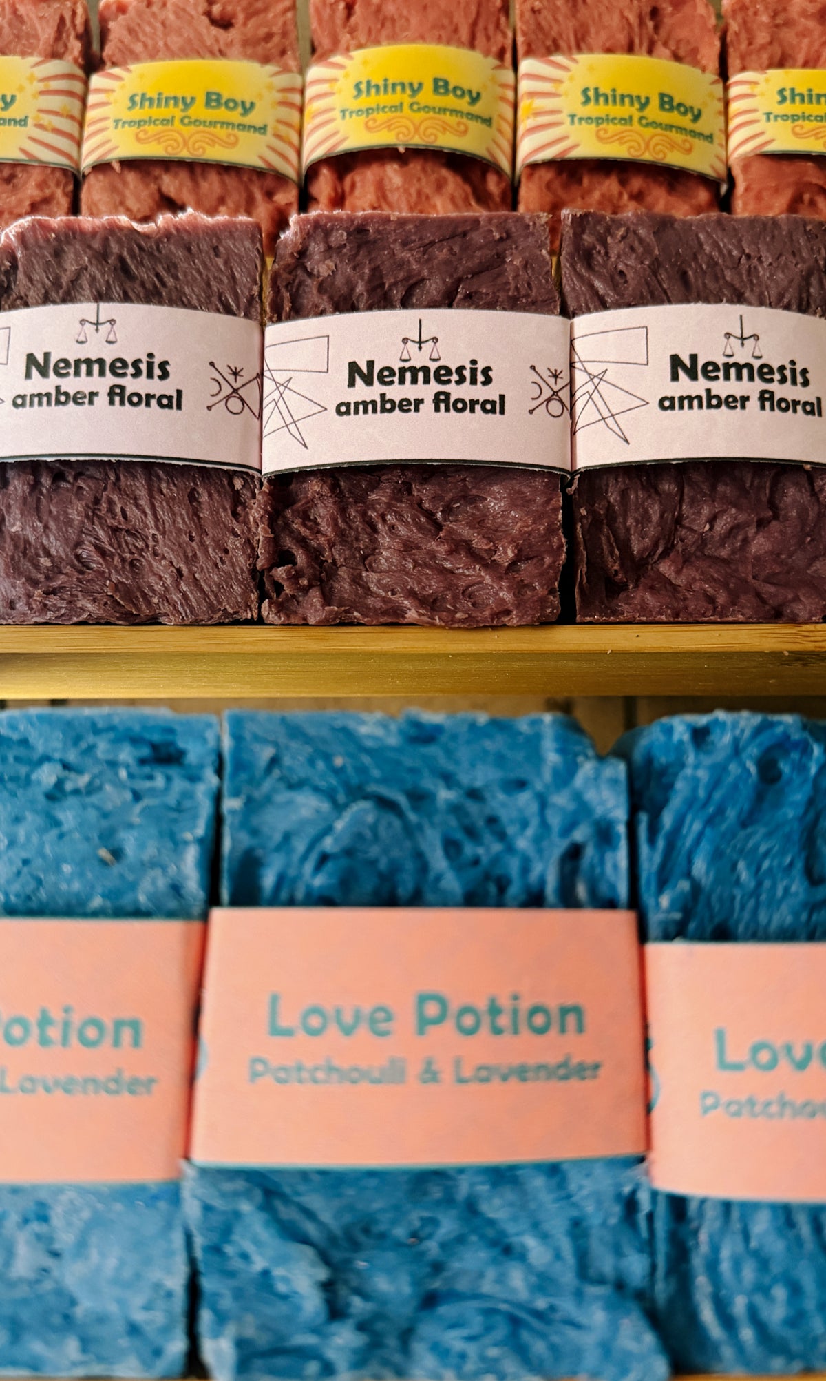 Bouncing Studio handmade bar soaps displayed on shelf including pink Shiny Boy Tropical Gourmand top row purple Nemesis Amber Floral middle row and blue Love Potion Patchouli and Lavender bottom row
