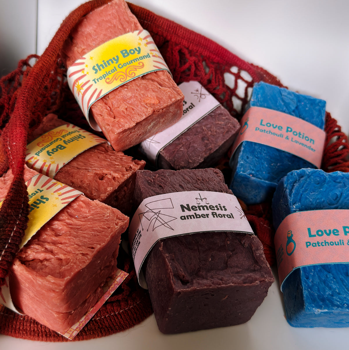 Assorted Bouncing Studio bars displayed in a crimson knit mesh bag: Shiny Boy (coral), Nemesis (maroon), and Love Potion (blue) bars visible with their branded labels