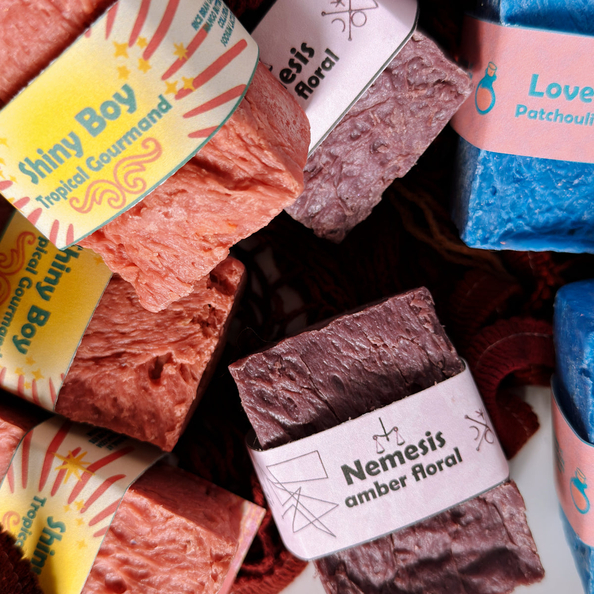Assorted Bouncing Soaps bars displayed in a crimson knit mesh bag: Shiny Boy (coral), Nemesis (maroon), and Love Potion (blue) bars visible with their branded labels