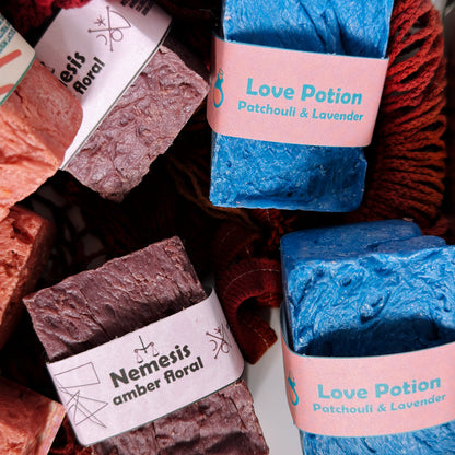 Assorted Bouncing Studio handmade bar soaps in a red rope basket including blue Love Potion Patchouli and Lavender and purple Nemesis Amber Floral