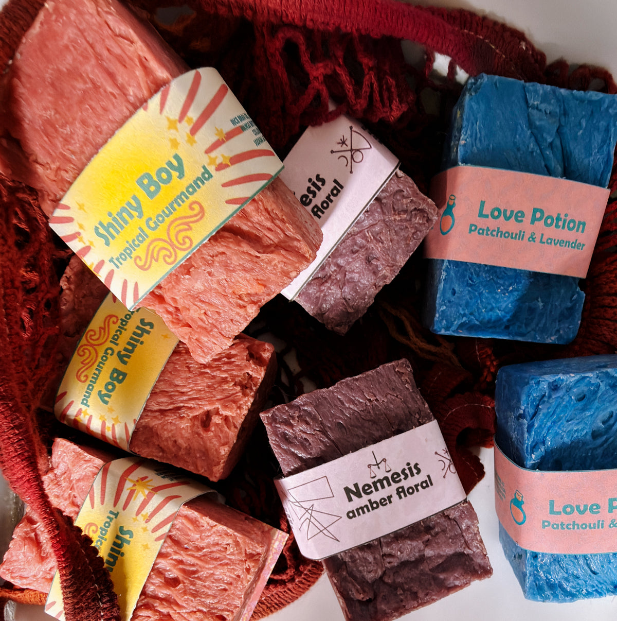 Assorted Bouncing Studio bars displayed in a crimson knit mesh bag: Shiny Boy (coral), Nemesis (maroon), and Love Potion (blue) bars visible with their branded labels