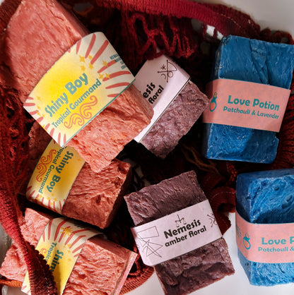 Assorted Bouncing Studio bars displayed in a crimson knit mesh bag: Shiny Boy (coral), Nemesis (maroon), and Love Potion (blue) bars visible with their branded labels