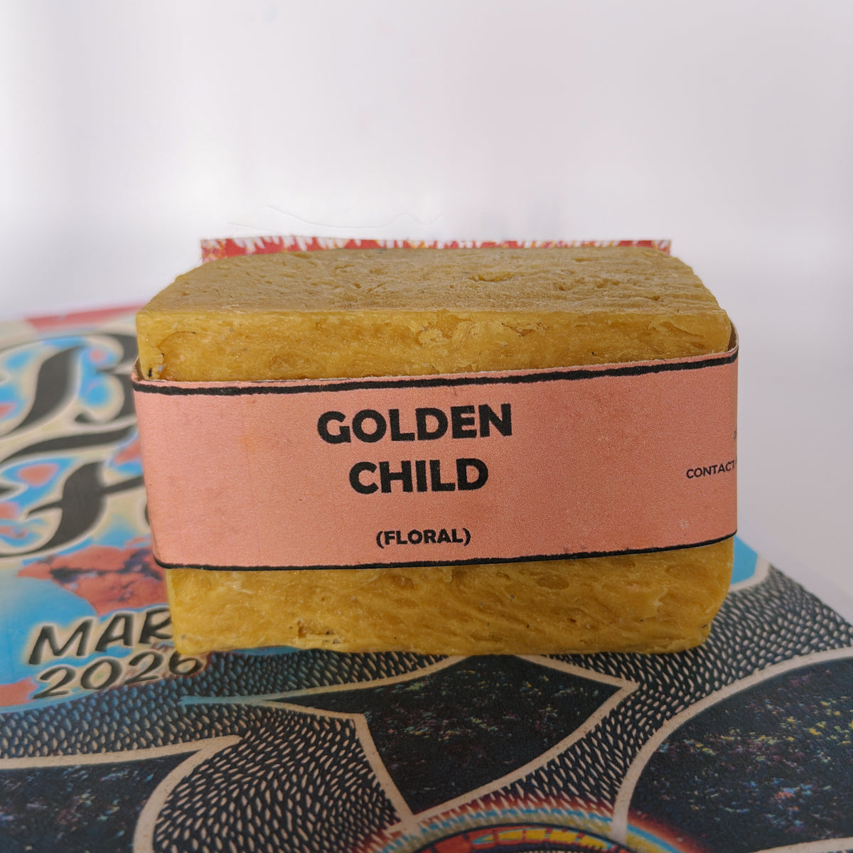 Single bar of Golden Child floral handmade soap by Bouncing Studio with golden yellow color and pink label resting on Bouncing Soaps branded booklet