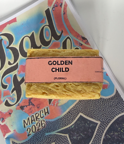 Single bar of Golden Child floral handmade soap by Bouncing Studio with golden yellow color and pink label resting on Bouncing Soaps branded booklet